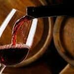 primo piano di bottiglia di vino rosso che viene versato in un calice sullo sfondo si intravedono due grosse botti