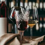 primopiano di 3 calici 2 con vini rossi e uno bianco un tovagliolo e una bottiglia di vino su un tavolino sullo sfondo c'è una scaffalatura piena di bottiglie di vino