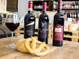 su un tavolo in legno in primo piano un tarallo appoggiato su una pagnotta con dietro tre bottiglie di vino rosso e sulla sinistra un bicchiere con del vino rosso