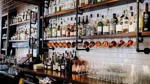 immagine di bancone e il dietro di un'enoteca stile industrial con bicchieri di vari modelli tazze in rame e scaffali con vari distillati sul bancone in fondo c'è anche un tablet per ordinazioni