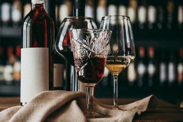 primopiano di 3 calici 2 con vini rossi e uno bianco un tovagliolo e una bottiglia di vino su un tavolino sullo sfondo c'è una scaffalatura piena di bottiglie di vino
